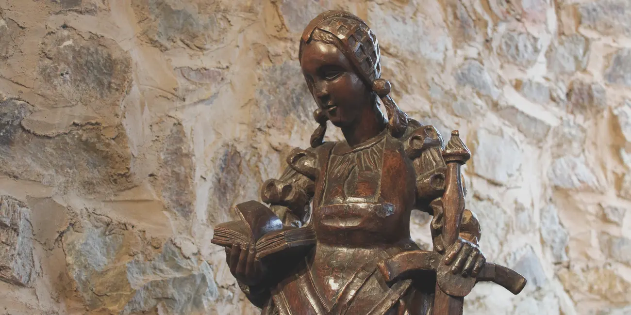 Statue de sainte Catherine d'Alexandrie en bois foncé vêtue d’une ample robe recouvrant une chemise plissée. Elle porte une coiffe réticulée d’où s’échappent deux nattes tressées. Dans la main droite, elle tient un livre qui symbolise son érudition et dans l’autre main une épée qui rappelle son martyre par décapitation.