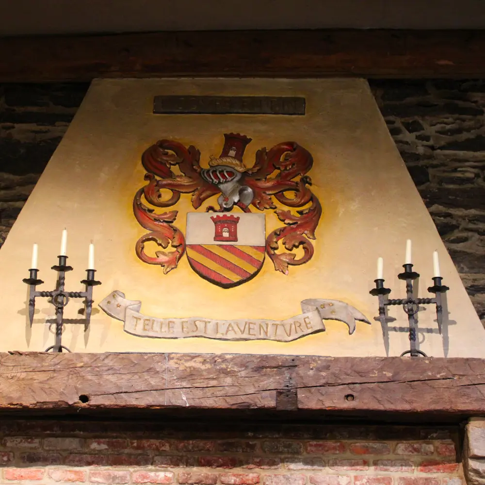 Hotte de la cheminée de la salle des Chevaliers du château de Reinhardstein portant en son centre le blason du professeur Jean Overloop avec son écu coiffé d’un heaume entouré de lambrequins, surmonté du cri de guerre de sa famille, JUSQUES EN FIN, et surplombant un listel arborant la devise familiale TELLE EST L’AVENTURE.