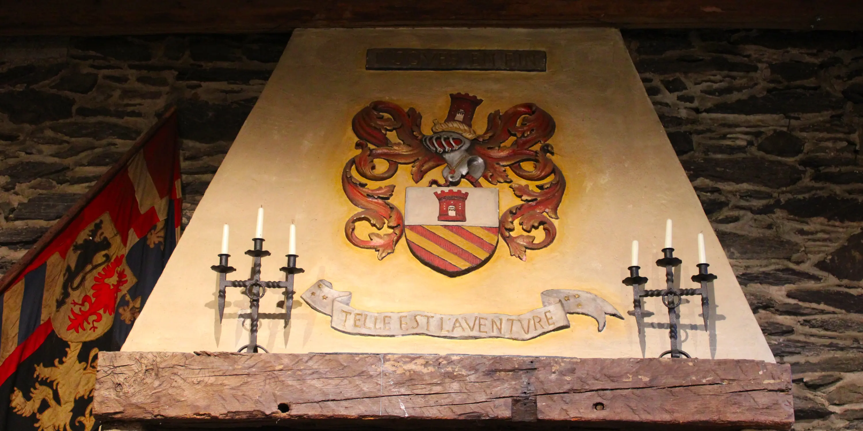 Hotte de la cheminée de la salle des Chevaliers du château de Reinhardstein portant en son centre le blason du professeur Jean Overloop avec son écu coiffé d’un heaume entouré de lambrequins, surmonté du cri de guerre de sa famille, JUSQUES EN FIN, et surplombant un listel arborant la devise familiale TELLE EST L’AVENTURE.