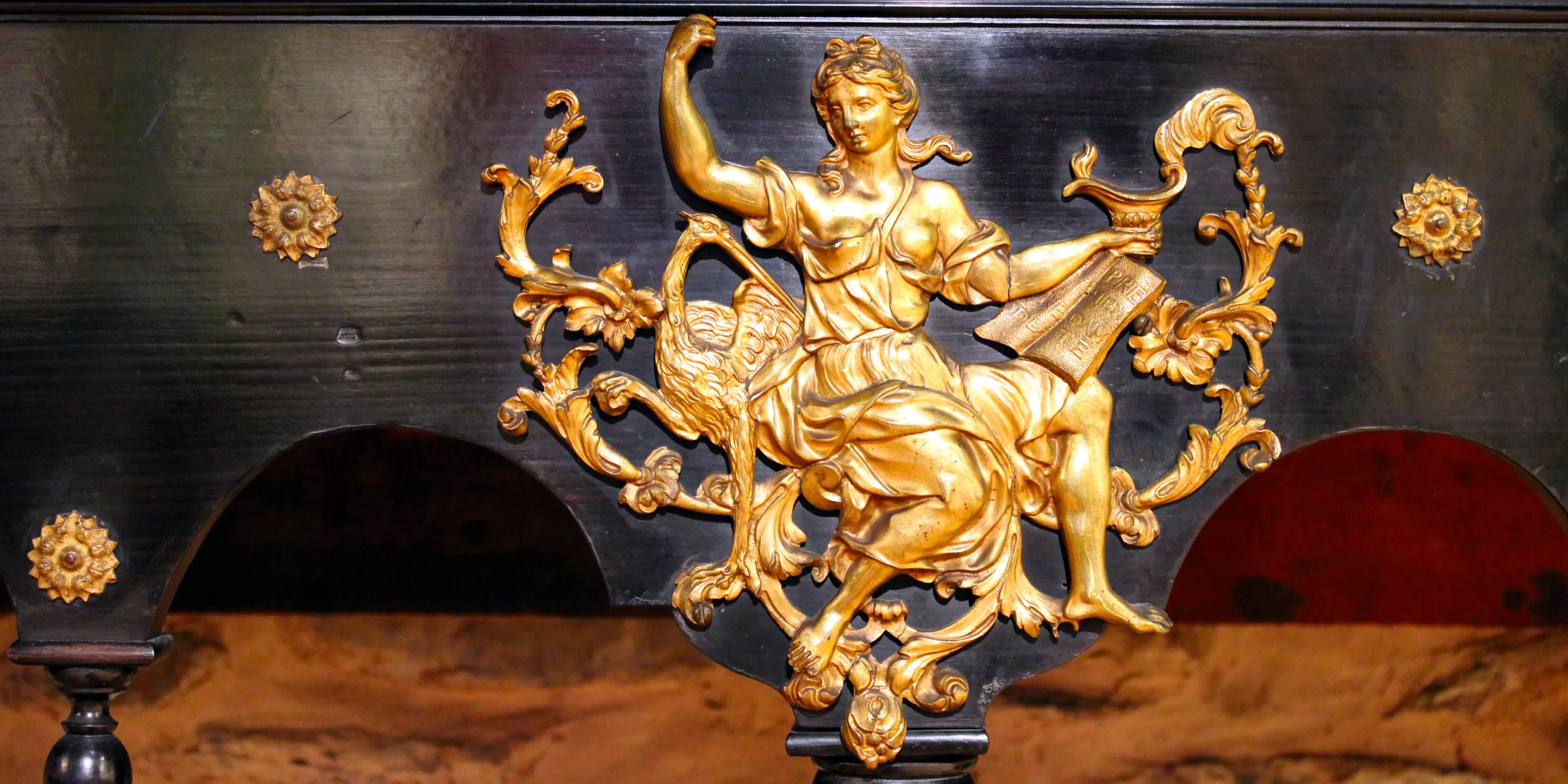 Ornement en bronze doré fixé sur un fond noir montrant une femme vêtue d'une longue robe à l'antique accompagnée d'une cigogne et tenant une lampe. Cet élément rehausse le piétement en bois noirci d'un cabinet anversois du XVIIe siècle. La déesse est Junon Lucine, déesse romaine de la naissance et des femmes.