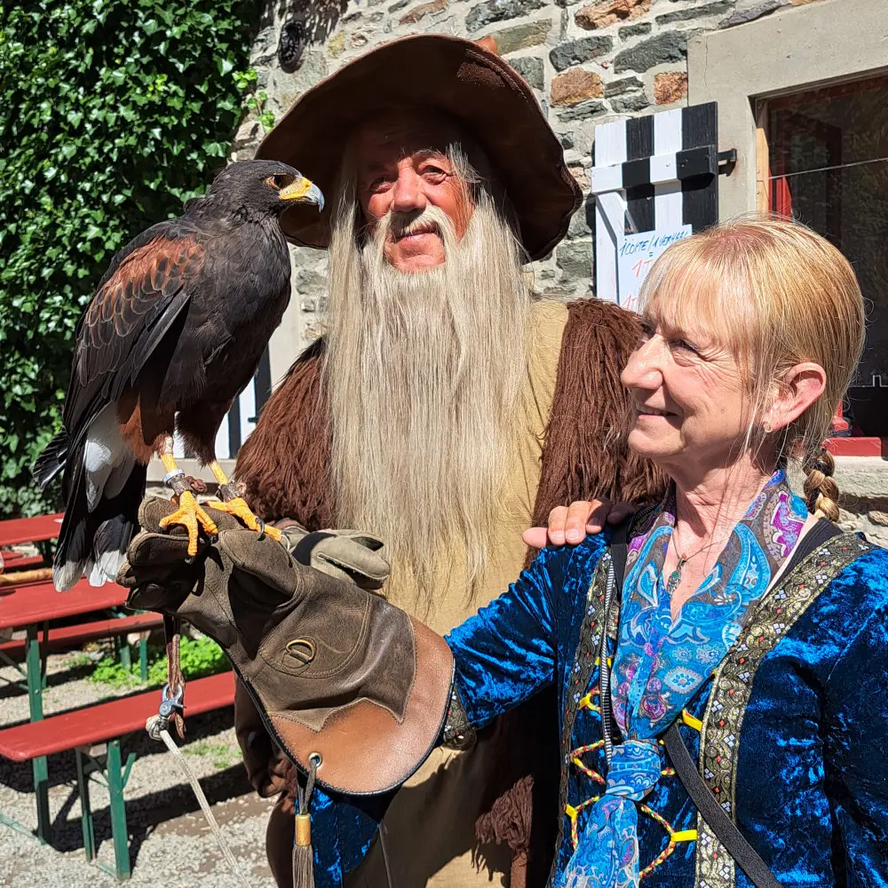 Fauconnier en habit du Moyen Âge portant sur sa main une buse de Harris devant la burghaus du château de Reinhardstein lors d’une animation médiévale, avec à sa droite Madame Andrée Deblond, guide au château de Reinhardstein, en tenue d’époque.