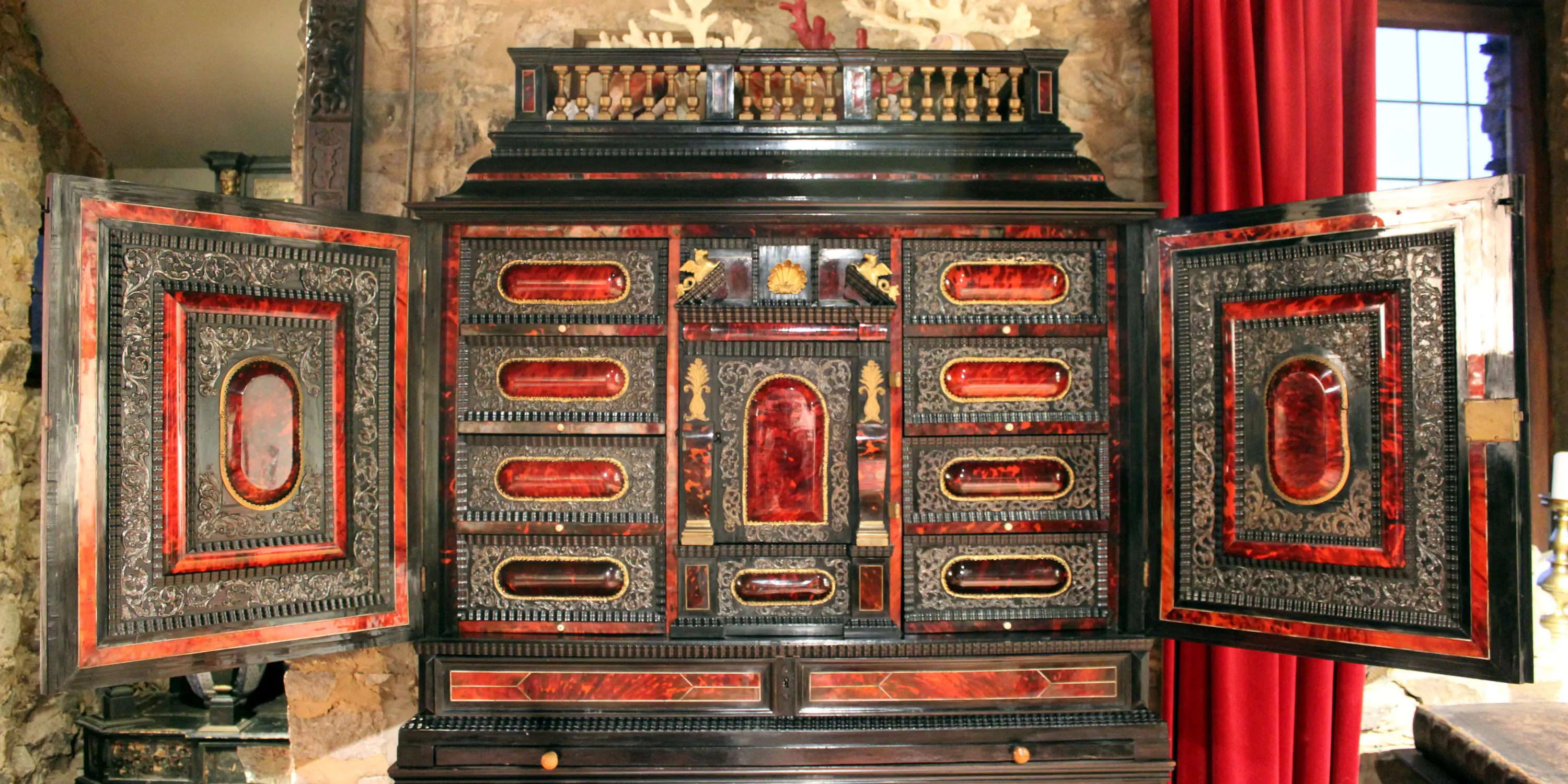 Cabinet anversois du XVIIe siècle en placage d’ébène enrichi de médaillons en écaille de tortue sur fond rouge qui sont encadrés de rinceaux d’argent repoussé ainsi que d’éléments en bronze doré qui enrichissent ce meuble d’apparat présenté ouvert, les vantaux laissant apparaître les nombreux tiroirs.