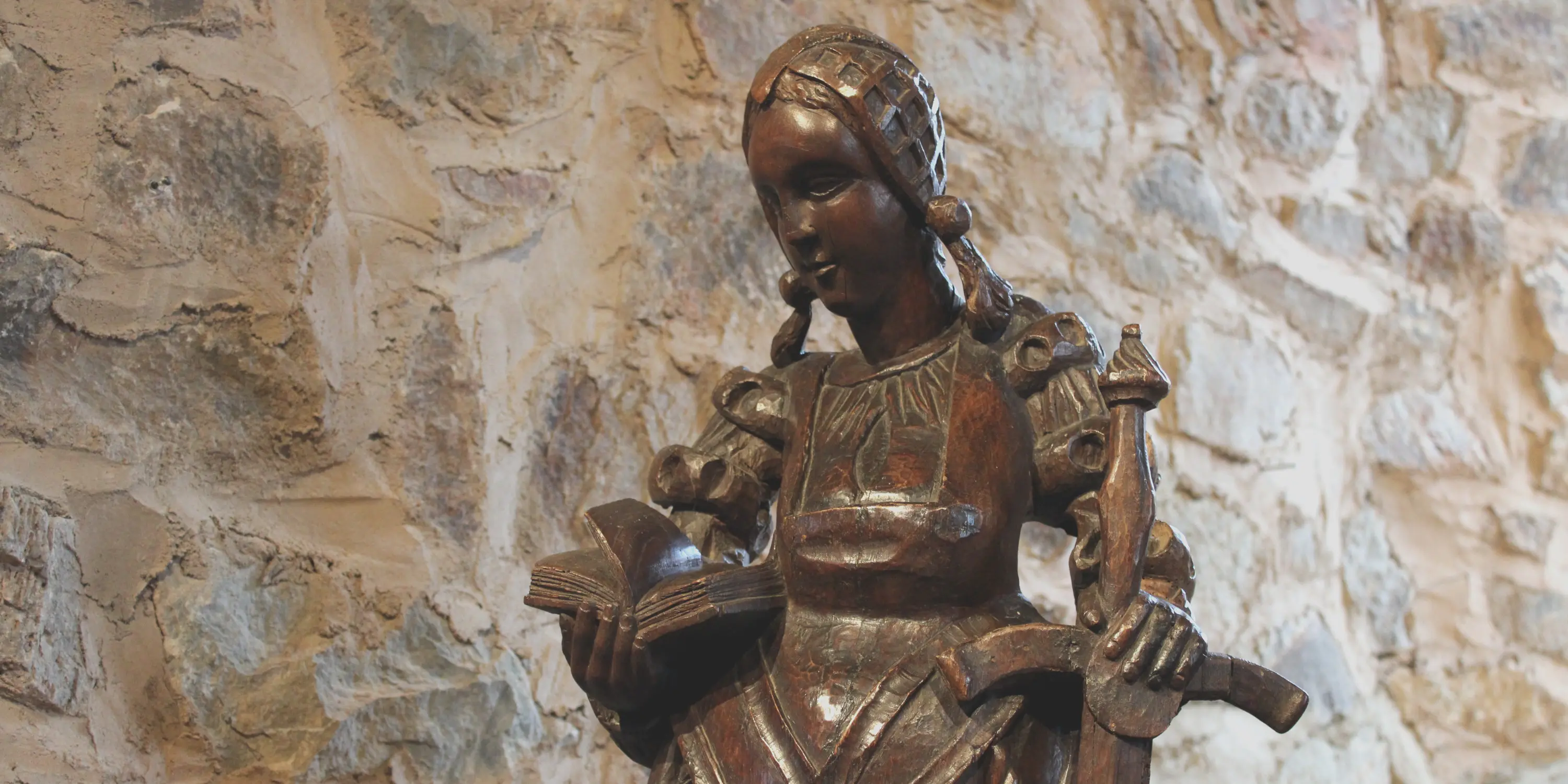 Statue de sainte Catherine d'Alexandrie en bois foncé vêtue d’une ample robe recouvrant une chemise plissée. Elle porte une coiffe réticulée d’où s’échappent deux nattes tressées. Dans la main droite, elle tient un livre qui symbolise son érudition et dans l’autre main une épée qui rappelle son martyre par décapitation.