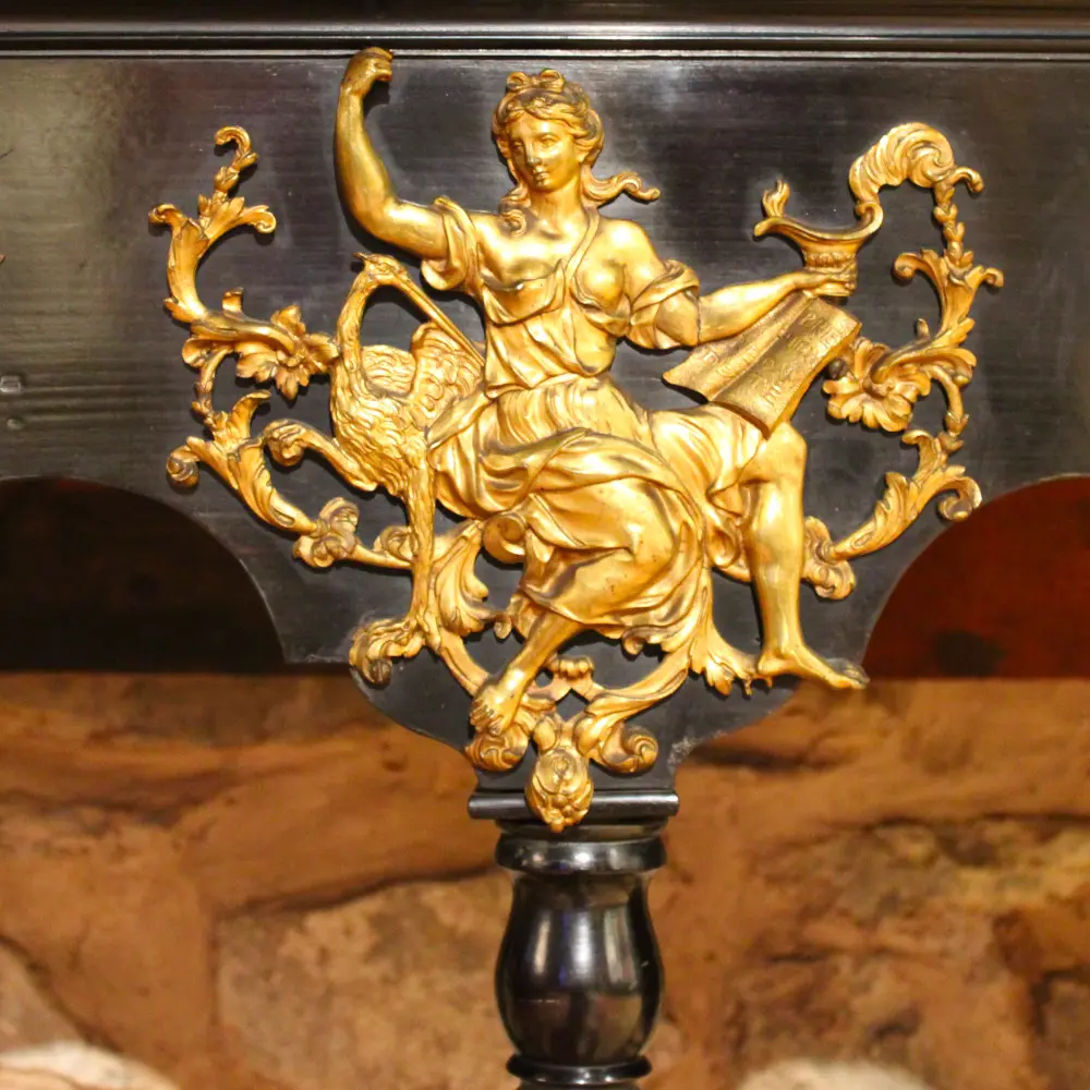 Ornement en bronze doré fixé sur un fond noir montrant une femme vêtue d'une longue robe à l'antique accompagnée d'une cigogne et tenant une lampe. Cet élément rehausse le piétement en bois noirci d'un cabinet anversois du XVIIe siècle. La déesse est Junon Lucine, déesse romaine de la naissance et des femmes.