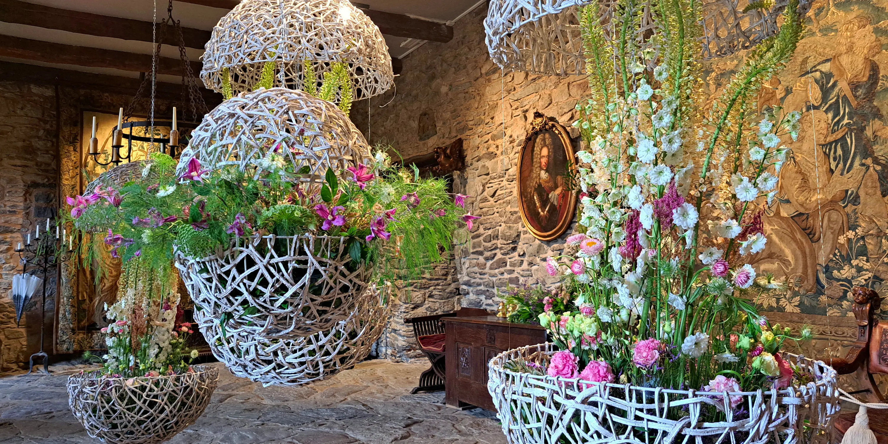 Delphiniums, clématites et œillets dans des cocons ajourés en osier blanchi suspendus par de fines chaînes métalliques ancrées aux poutres du plafond de la salle des Chevaliers du château de Reinhardstein lors de l’événement Castelflora.