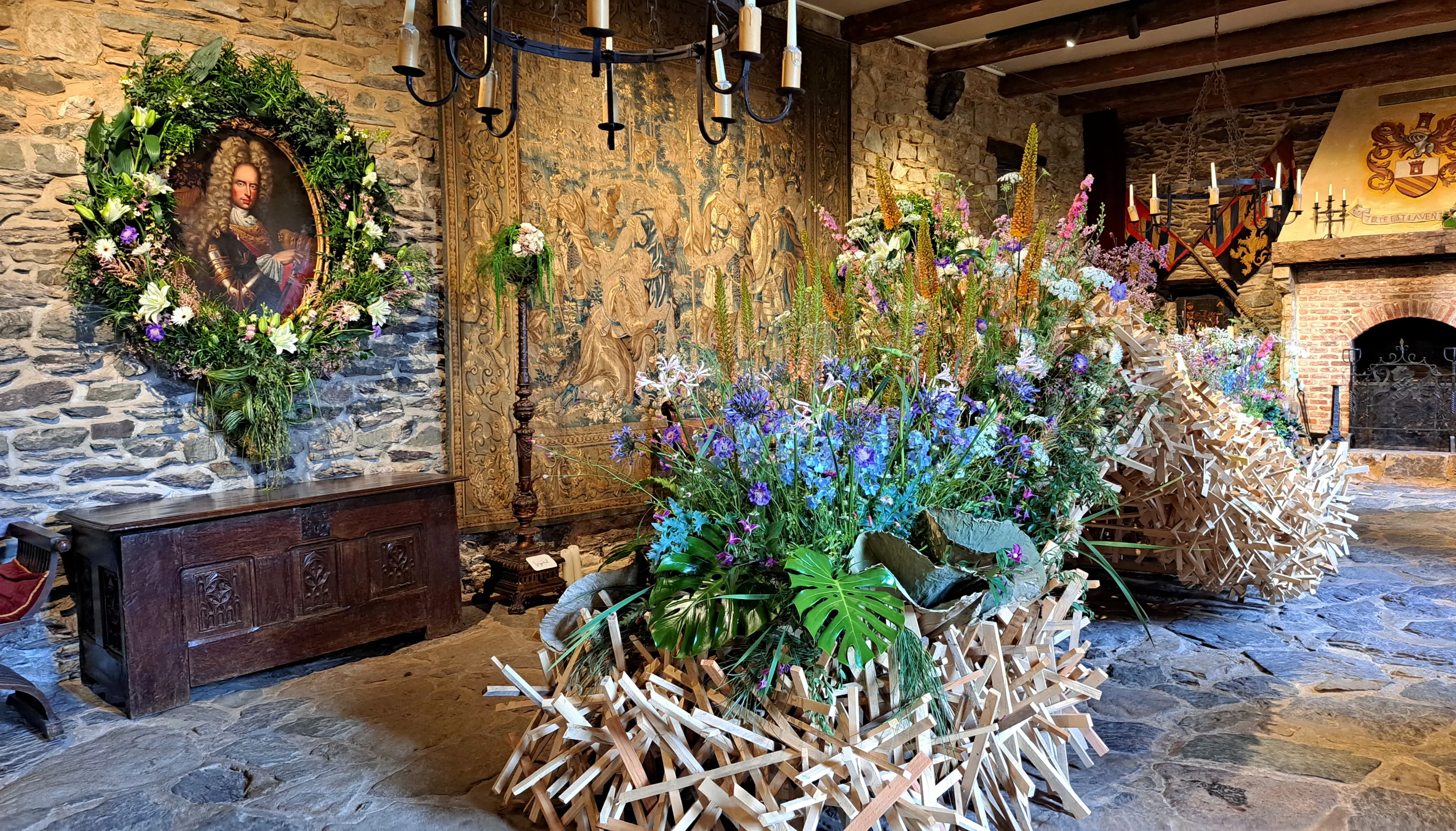 Delphiniums, lis des steppes, agapanthes et alliums mis en valeur par un feuillage de fougères, de graminées et de monstera deliciosa dans la salle des Chevaliers du château de Reinhardstein, lors de l’événement Castelflora, où le portrait de Joseph Ier de Habsbourg est encadré par une couronne florale de lys blancs et rosés.