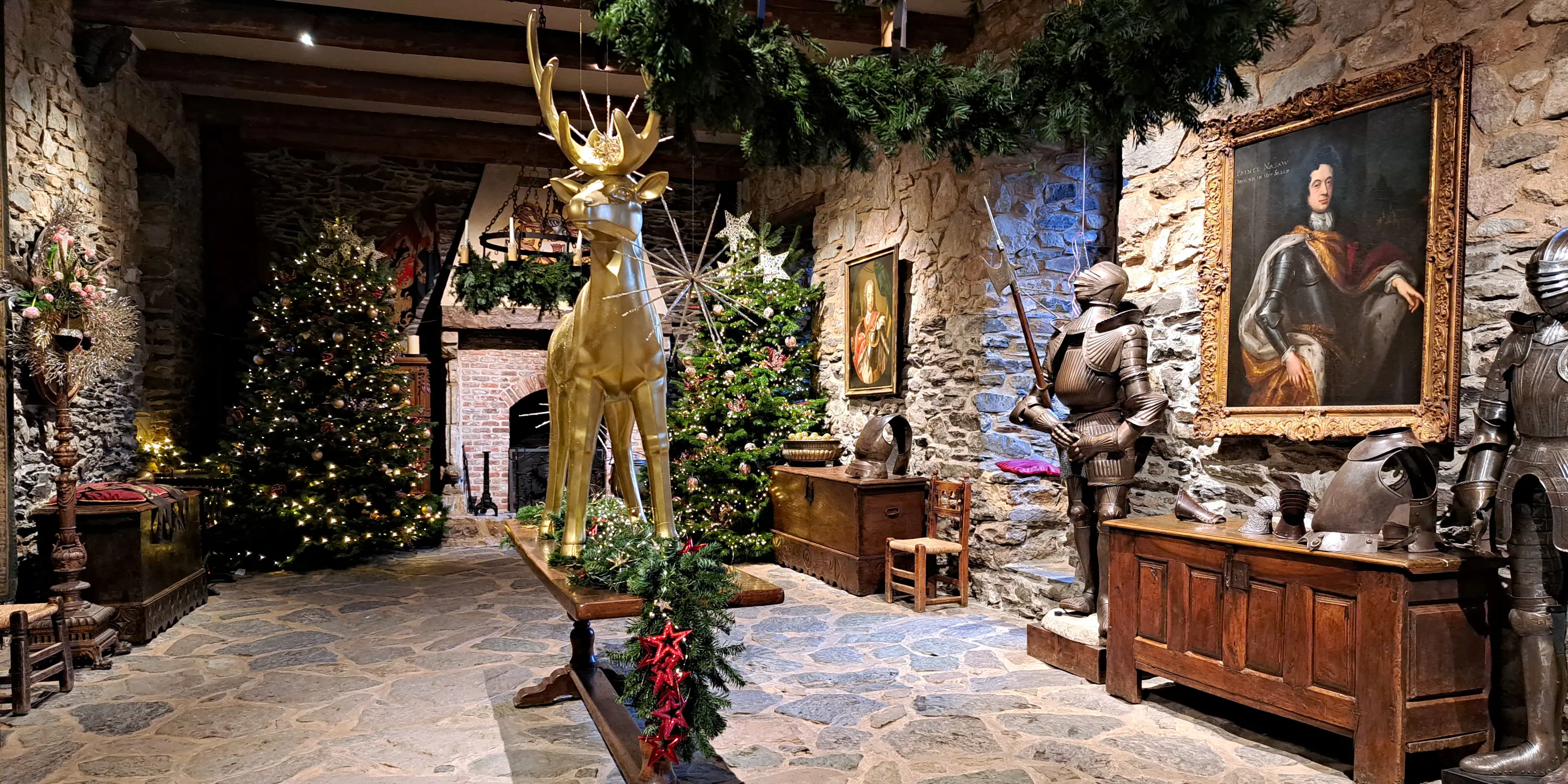 Décoration de la salle des chevaliers du château de Reinhardstein, le 24 décembre 2022, lors de l’événement Noël au Château comprenant une statue de renne doré posée sur une table en chêne, deux armures du XIXe siècle, deux tableaux, deux coffres ainsi que des guirlandes et des étoiles suspendues à deux sapins de Noël.